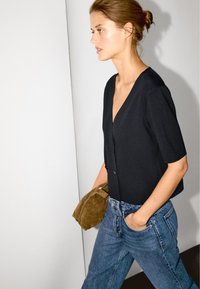 Schwarze kurzärmelige Strickjacke mit V-Ausschnitt und zwei Knöpfen, kombiniert mit hoch taillierten blauen Denim-Jeans. Das Model trägt eine braune Wildledertasche.