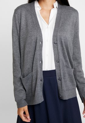 Femme portant un cardigan gris boutonné sur une chemise blanche et une jupe plissée bleu marine, debout devant un fond uni.