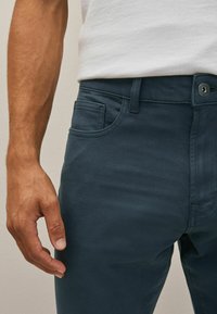 Chino dark teal cu o croială slim, dotat cu un buzunar frontal, curea pentru brâu și închidere cu un buton metalic. Textură de țesătură moale.