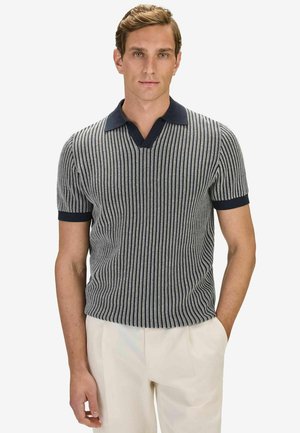 Man som bär en svart och vit pikétröja med korta ärmar och vertikala ränder, med navyblå krage och vita byxor, stående med ena handen i fickan.