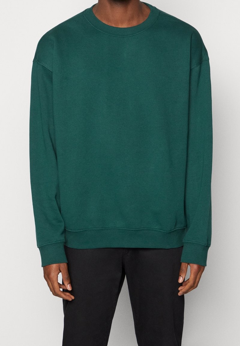 Homme portant un sweat-shirt à manches longues vert foncé uni associé à un pantalon noir, debout devant un fond clair.