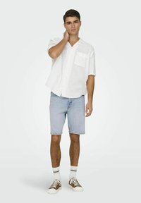 Cămașă albă cu mânecă scurtă și buzunar, șorturi din denim de culoare albastră deschisă și adidași maro cu talpă albă. Modelul stă într-un cadru luminos.