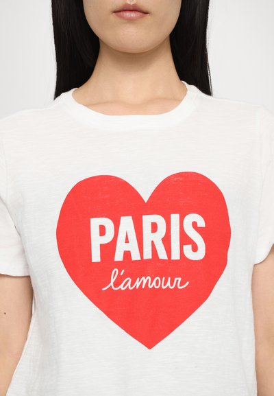 Valkoinen puuvillapaita, jossa on iso punainen sydänkuvio ja "PARIS" paksuilla valkoisilla kirjaimilla sekä "l'amour" kursiivilla alla.