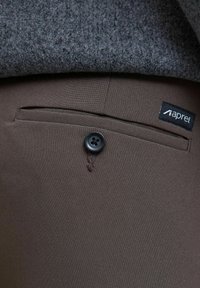 Pantalons en tissu marron avec une poche latérale, dotés d'un bouton noir et d'une petite étiquette de marque étiquetée "aprel". Couche en tricot gris texturé au-dessus.