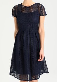 Laivansinis mekko pitsiyksityiskohdalla, lyhyet hihat, tiukka vartalo-osa ja levenevä hame. Sisältää geometrisia kuvioita ja polvenpituinen helma.