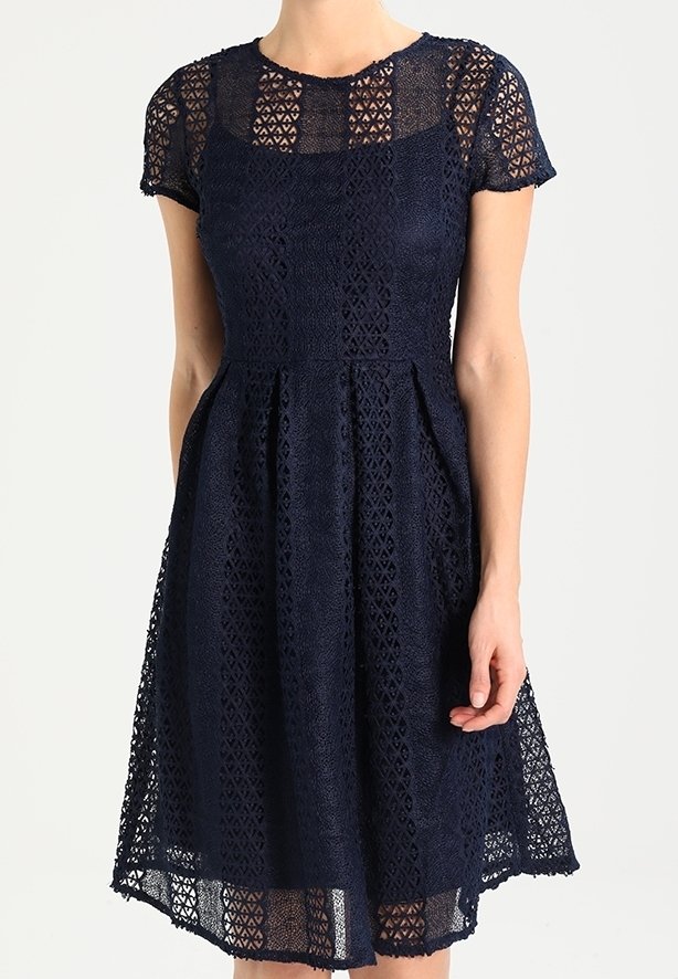 Laivansinis mekko pitsiyksityiskohdalla, lyhyet hihat, tiukka vartalo-osa ja levenevä hame. Sisältää geometrisia kuvioita ja polvenpituinen helma.