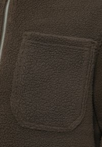Chaqueta de forro polar marrón con una superficie texturizada, un bolsillo cuadrado prominente y una cremallera plateada. Material suave con un aspecto acogedor.