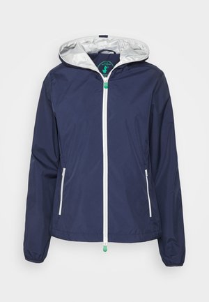 Veste légère bleu marine avec une capuche doublée d'argent, une fermeture éclair blanche et des poches latérales. Elle possède des poignets élastiques et un ourlet à cordon de serrage.