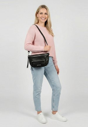 Lachende vrouw in een roze trui, lichtblauwe spijkerbroek, witte sneakers, met een zwarte crossbodytas met ritszakken tegen een grijze achtergrond.