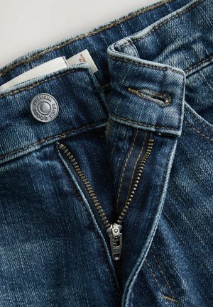 Jeans en denim bleu foncé avec une fermeture éclair, une fermeture éclair en laiton et un bouton. Présente des coutures visibles et une texture légèrement usée.