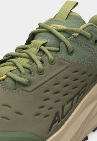 Κοντινό πλάνο πράσινου και μπεζ παπουτσιού για τρέξιμο σε μονοπάτι, με υφή δίχτυ, παχύ μαξιλαρωμένο πέλμα, κορδόνια και σήμανση "ALTRA" στο πλάι.