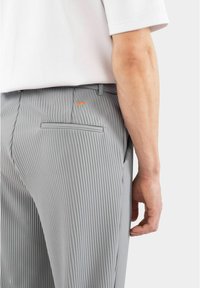 Graue gestreifte Hosen mit schmaler Passform, ausgestattet mit einer Gesäßtasche und einem orangefarbenen Akzent in der Nähe des Bundes. Glatte Stofftextur und maßgeschneiderter Schnitt.
