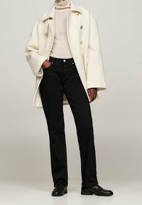 Manteau croisé en mélange de laine crème avec des boutons dorés, associé à un jean noir classique. Texture douce et coupe décontractée. Mocassins noirs.