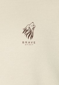 Lichtbeige sweater met een bruine wolfgrafiek en de tekst "BRAVE SPIRIT." Zachte textuur met een ronde hals.