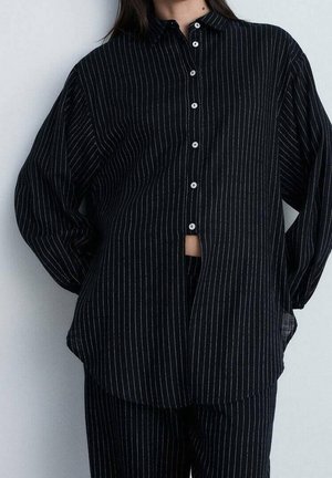 Woman wearing black long-sleeve button-up shirt and matching pants with thin white vertical stripes, standing with hands behind back.