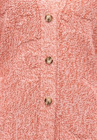 Κοντινό πλάνο πορτοκαλί και λευκής πλεκτής ζακέτας με γεωμετρικά μοτίβα και μπεζ στρογγυλά κουμπιά κατά μήκος του κέντρου.