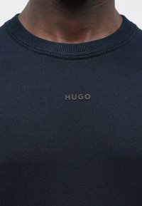 Meresinine kampsun, silmusega kaelusega, tekstureeritud kangaga, kus on rinnal must "HUGO" logo.