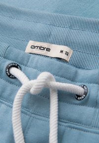 Lyseblå joggingbukser med hvid snøre bundet i en knude, der viser "ombre" mærkelabel og størrelse M (EUR 48) på taljebåndet.