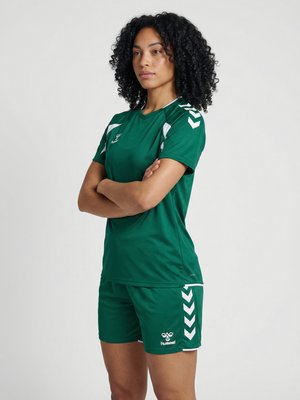 Vrouw met krullend haar staat met arms over elkaar, draagt groene sportkleding met witte chevron-details op mouwen en short.