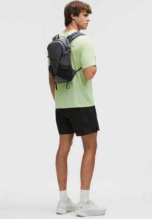 Musta ja harmaa reppu, jossa säädettävät hihnat, valmistettu kestävästä materiaalista, pidetty vaaleanvihreän paidan päällä, yhdistetty mustiin shortseihin ja valkoisiin kenkiin.