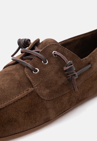 Chaussure mocassin en daim marron avec lacet en cuir passé à travers des œillets métalliques et détails en cuir noué sur un fond clair.