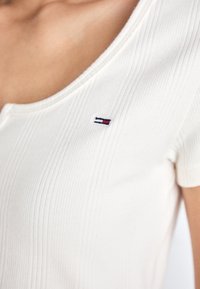 Biała, prążkowana, krótko rękawowa bluzka z dekoltem w serek oraz małym, haftowanym logo w kolorach czerwonym, niebieskim i białym na lewej piersi.