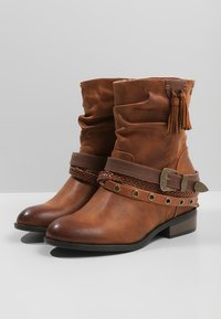 Stivali alla caviglia in pelle marrone con un design morbido, dettagli con fascia intrecciata, borchie metalliche e nappine sui lati. Tacco piatto.