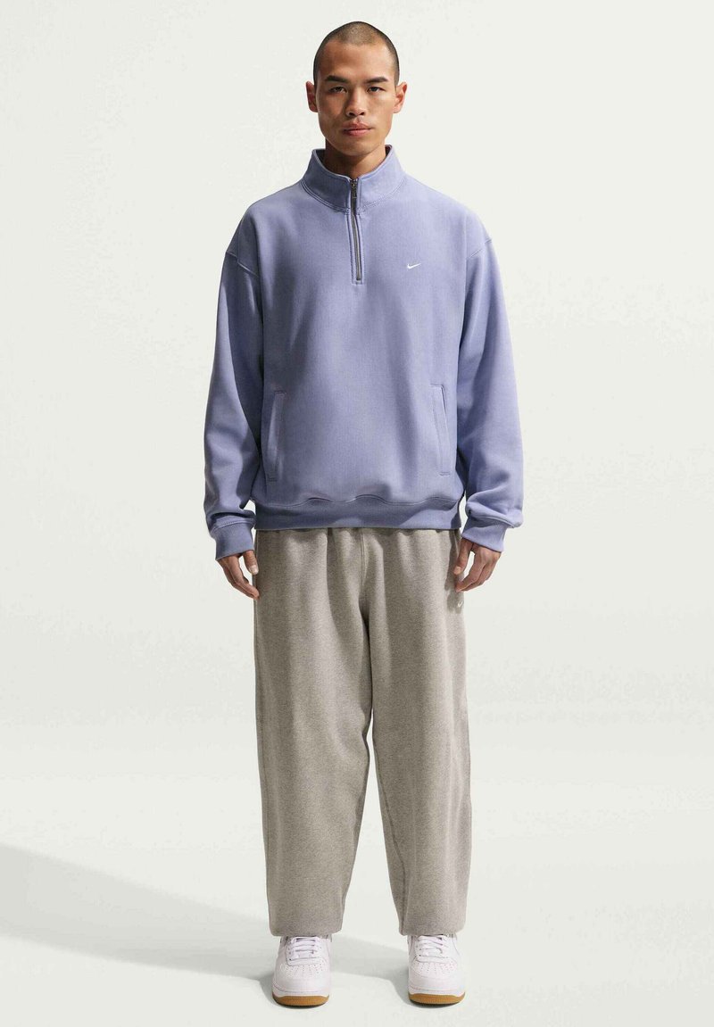 Sweat-shirt à fermeture éclair de couleur violet clair avec manches longues et poches latérales, associé à un pantalon de survêtement gris et des baskets blanches. Textures lisses et douces.