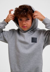 Grå bomulls-hoodie med stor huva och en logotypmärke på bröstet. Mjuk textur, avslappnad passform med ribbade muddar och fåll.