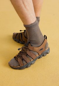 Brahns ādas sandales ar acs sietu akcentiem, aprīkotas ar regulējamu saites sistēmu un gumijas zoli. Valkā ar pelēkiem vītnes zeķēm.