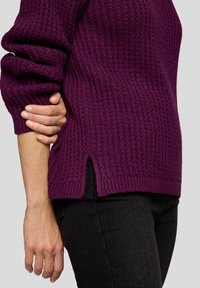 Lila gestrickter Pullover mit einem strukturierten Muster, ausgestattet mit umgeschlagenen Bündchen und seitlichen Schlitzen am Saum, kombiniert mit schwarzen Jeans.