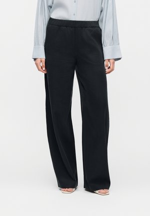 Kvinde iført sorte bukser med vide ben og elastisk talje, lyseblå langærmet bluse og beige åbne sandaler, stående på ensfarvet baggrund.