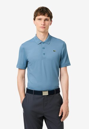 Junger Mann, der ein hellblaues kurzärmeliges Poloshirt mit einem kleinen grünen Krokodil-Logo und dunkle Hosen mit schwarzem Gürtel trägt.
