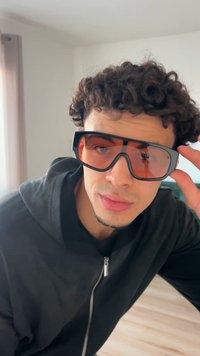 Γυαλιά ηλίου με χοντρό μαύρο πλαίσιο και μεγάλα, πορτοκαλί σκουρόχρωμα φακούς. Ο σχεδιασμός έχει αιχμηρές γωνίες και φαρδιές πλευρικές βάσεις.