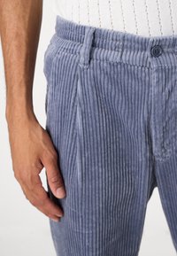 Šedé manšestrové kalhoty s vertikální žebrovanou texturou, s knoflíkovým zapínáním a postranními kapsami. Zobrazené vedle ruky pro měřítko.