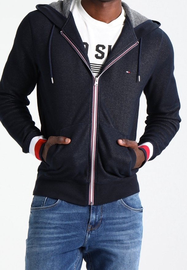 Homme portant un sweat à capuche bleu marine zippé avec des poignets rayés rouge, blanc et bleu et les mains dans les poches avant, associé à un jean bleu.