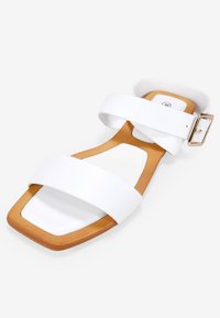 Sandal planas blancas con dos correas anchas, punta cuadrada y suela color tostado con un acento de hebilla metálica. Textura del material suave.