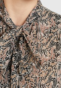 Blusa con colletto a farfalla, realizzata in tessuto leggero. Presenta una base crema con motivi astratti neri, marroni e arancioni.