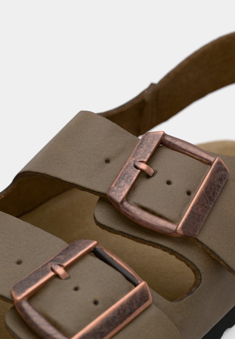 Sandale en cuir vegan marron avec deux larges sangles réglables dotées de boucles en cuivre texturé. Semelle intérieure douce beige au design minimaliste.