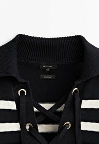 Pull en tricot noir à rayures horizontales blanches, col côtelé et devant à lacets avec œillets métalliques.