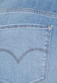 Tela de mezclilla azul claro con un bolsillo trasero que presenta un diseño de costura curva y un hilo marrón en contraste. Textura suave, sin patrones.