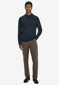 Mies, joka seisoo yhdellä kädellä taskussa pukeutuneena tummansiniseen pitkähihaiseen pikeepaitaan, ruskeisiin housuihin ja tummanruskeisiin kenkiin yksinkertaista taustaa vasten.