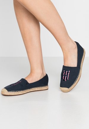 Person iført sorte slip-on espadrillos med flettede såler og broderede initialer i rød, hvid og blå på forsiden.