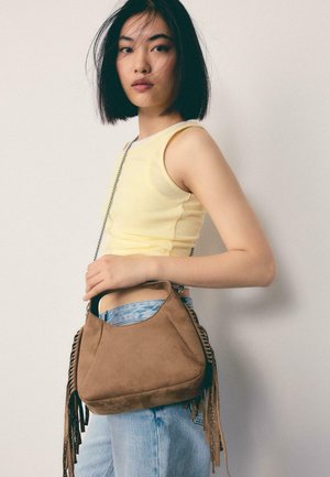 Mlada ženska s črnimi krajšimi lasmi nosi rumeno brezrokavnik in svetlo modre kavbojke, preko ramena drži rjavo torbico iz semiša z resicami.