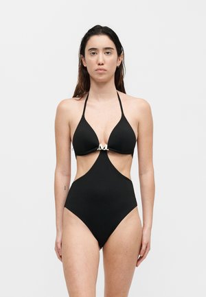 Γυναίκα που φοράει μαύρο ολόσωμο μαγιό με κοψίματα, λαιμό halter και μεταλλικό κούμπωμα μπροστά, στέκεται μπροστά σε απλό ανοιχτό φόντο.