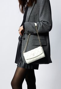 Sac à main matelassé blanc avec une chaîne dorée, doté d'une fermeture rabat et d'un logo en relief, posé à côté d'un blazer gris et d'une jupe plissée noire.