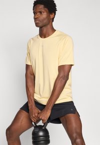 Κίτρινη αθλητική κοντομάνικη μπλούζα με στρογγυλή λαιμόκοψη και λογότυπο, συνδυασμένη με μαύρο σορτς. Το άτομο κρατάει μαύρο kettlebell.