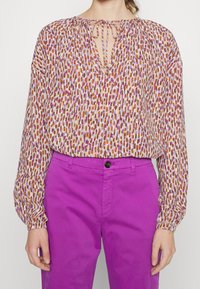 La blouse à manches longues présente un motif abstrait multicolore dans des tons de violet, orange et marron, avec un col à nouer et des détails froncés.