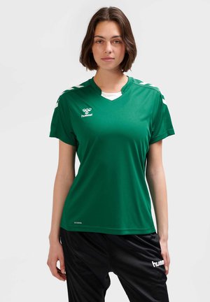 Kobieta o krótkich brązowych włosach, ubrana w zieloną koszulkę sportową hummel i czarne sportowe spodnie hummel na jednolitym tle.