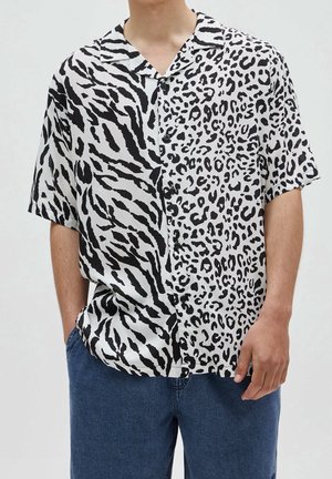 Homme portant une chemise à manches courtes boutonnée avec des rayures de tigre noires et blanches sur la moitié gauche et des taches de léopard sur la moitié droite, associée à un jean bleu.